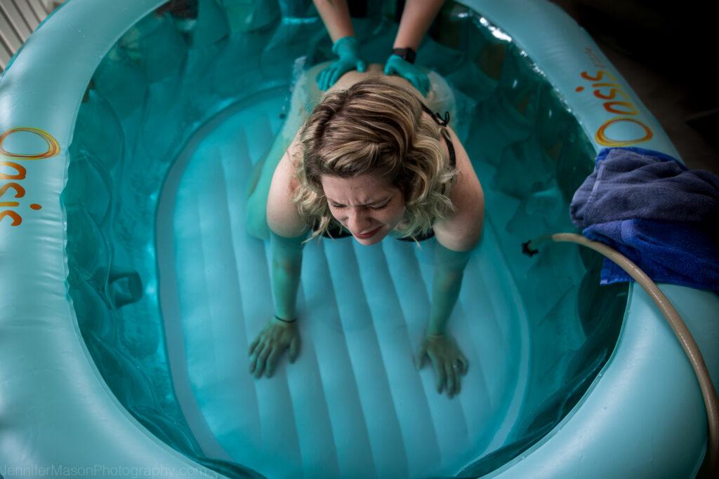 Blue water birth tub with a pregnant person in a hands and knees position. Gloved hands of a doula press on the back.