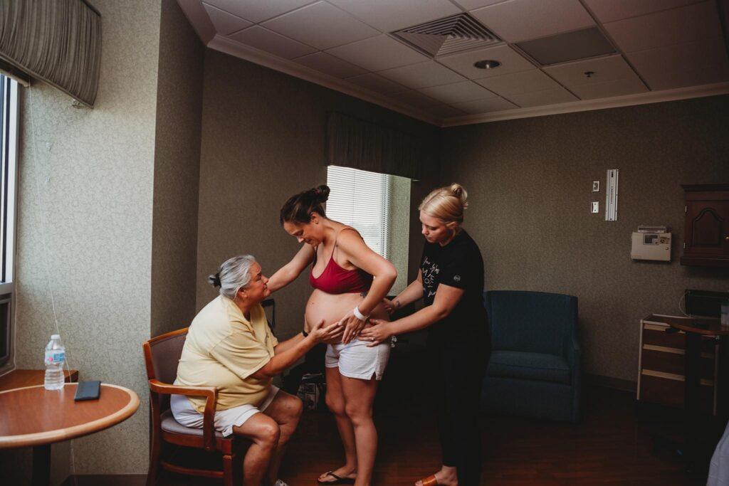 Doula in a black shirt squeezing the hips of a pegnant person in a red bra and white shorts.