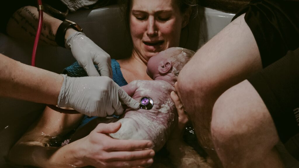 A brand new baby on the birthing parent's chest with hands from support people around.