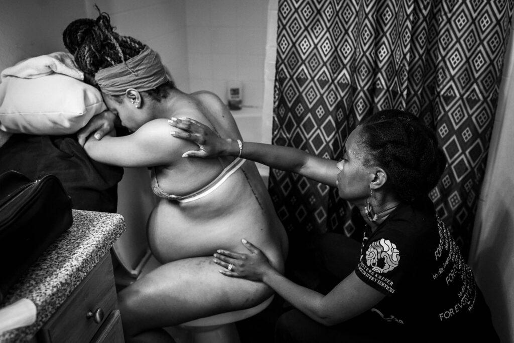 Doula massaging a client in labor's back while they sit backwards on a toilet.