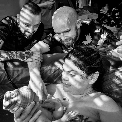 A woman gives birth in a water pool, assisted by two doulas, one holding a newborn baby; all show focused expressions in a dimly lit room.
