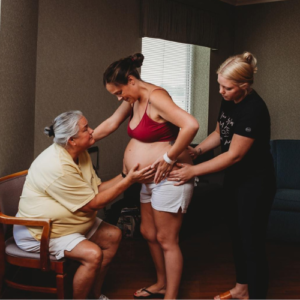 Doula in a black shirt squeezing the hips of a pregnant person in a red bra and white shorts.