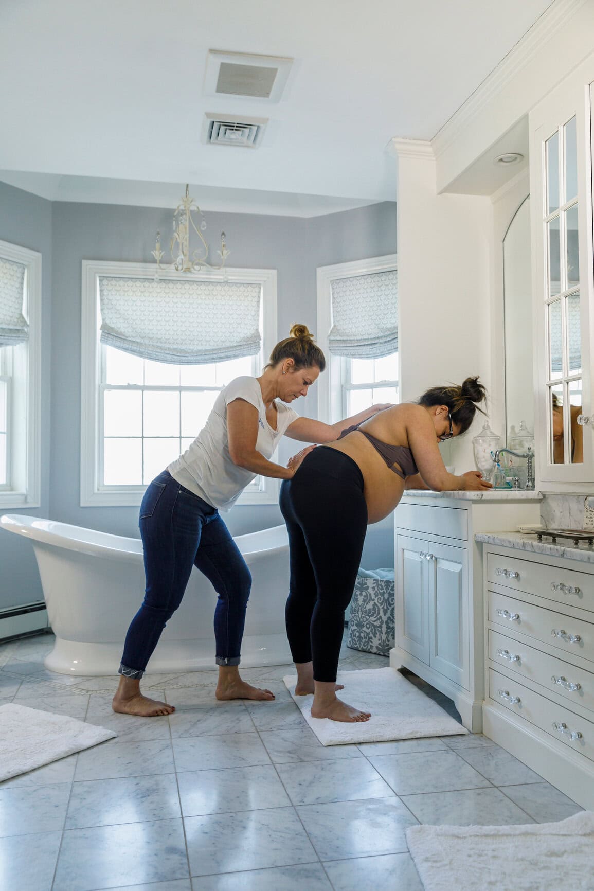 A doula applies hip pressure while a laboring person leans on a bathroom counter
